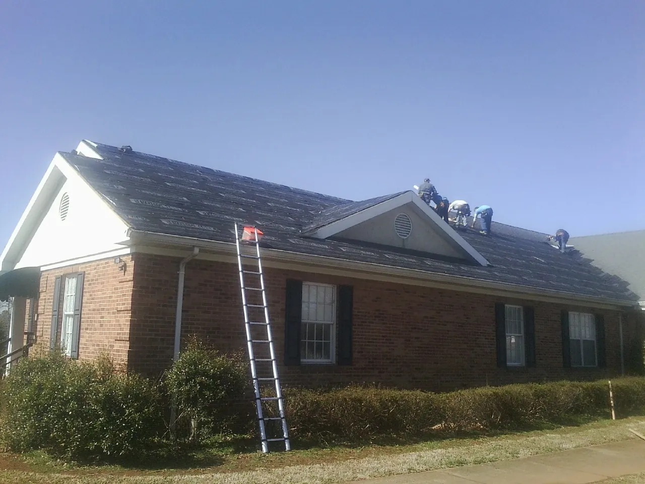 Crew tearing shingles off a brick ranch roof with a ladder set — Emergency Roof Repair work by our crew in Millican