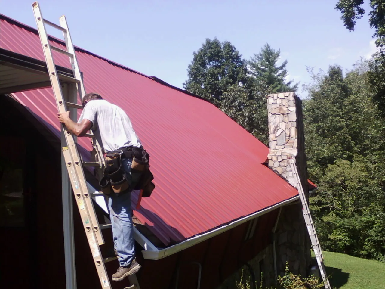 Roofer on a ladder beside a red metal roof and stone chimney — Soffit & Fascia Repair by Roofing Brothers in Millican